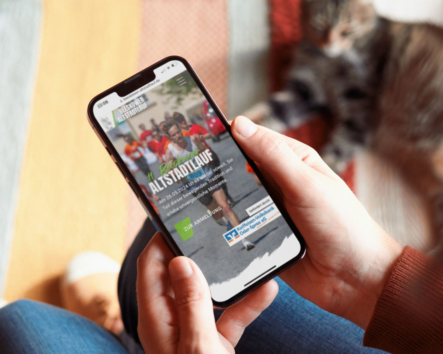 Eine Person hält ein Smartphone, auf dem eine Webseite für einen Stadtlauf angezeigt wird, während eine Katze im Hintergrund liegt.